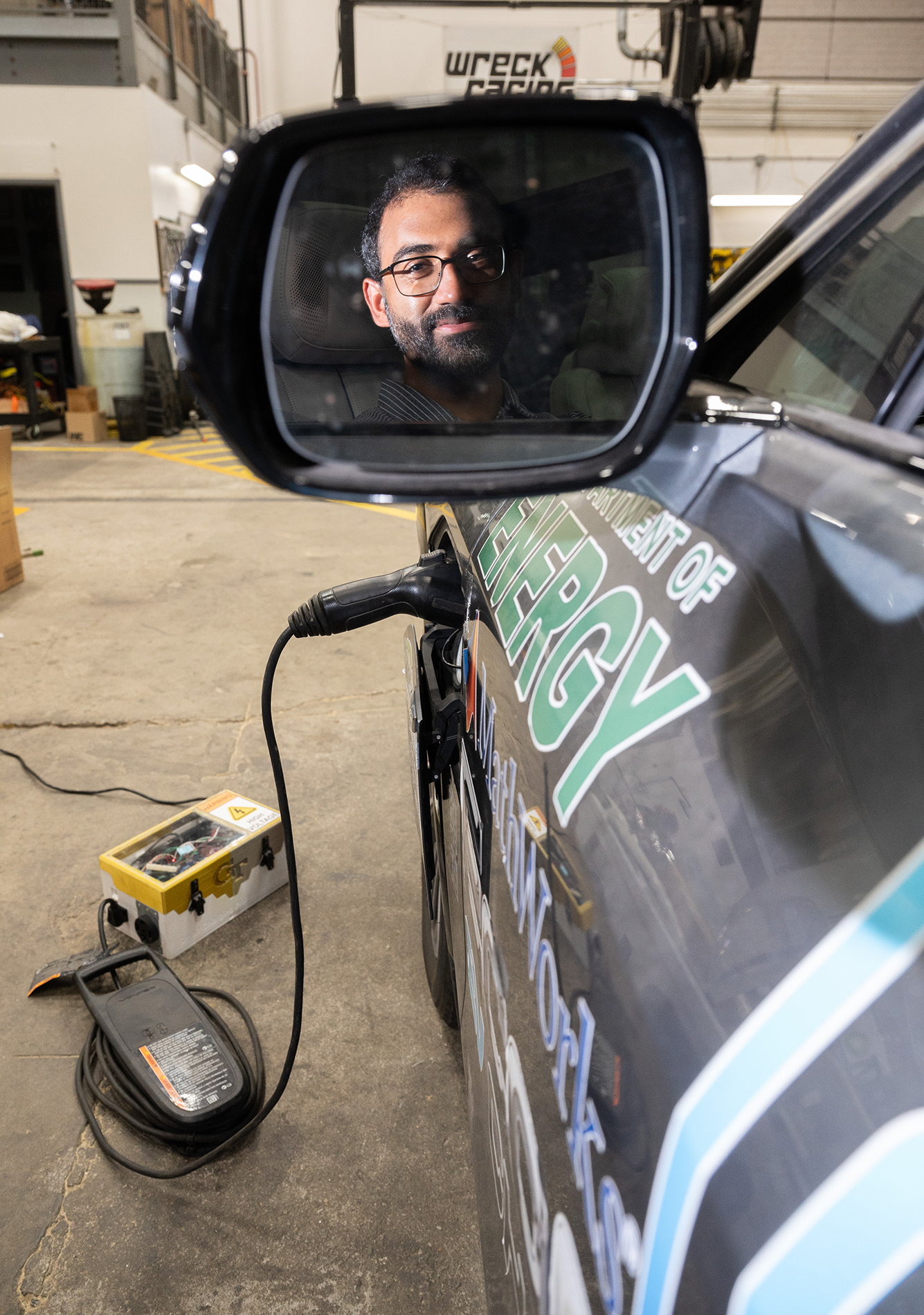 Male researcher's face shown in vehicle's side mirror while the electric vehicle is attached to a smart charger