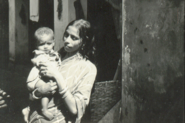 black and white image of a young Indian woman holding an infant while standing in front of a house