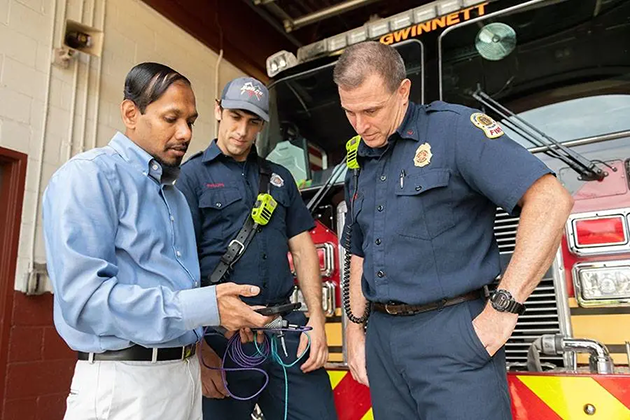 Man wearing a blue dress shirt with two firefighters in front of a fire truck while all three men look at a cellphone screen