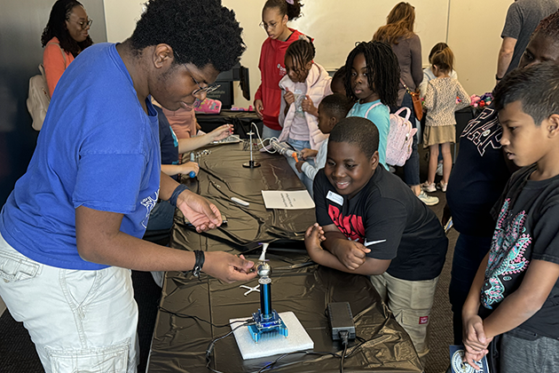 A room full of elementary school children watch two college graduate students conduct demonstrations on a table.
