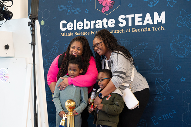 Two females and their children posing in front of a Celebrate STEAM background photo booth