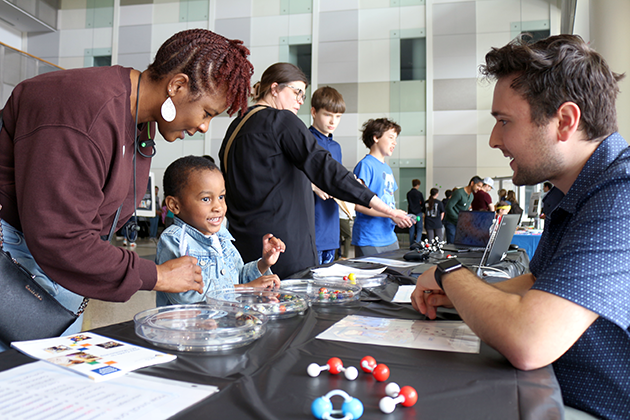 A mom and her son on one side of a table talk to a male graduate student about the scientific demo he is showing.