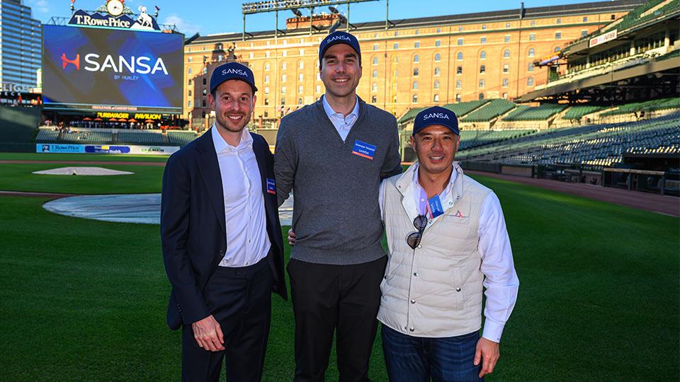 Three men who co-founded and lead Huxley Medical stand together on the field of a baseball stadium, smiling at the camera. They are wearing SANSA-branded hats, with a large video board behind them displaying the SANSA logo and the stadium seating in the background.