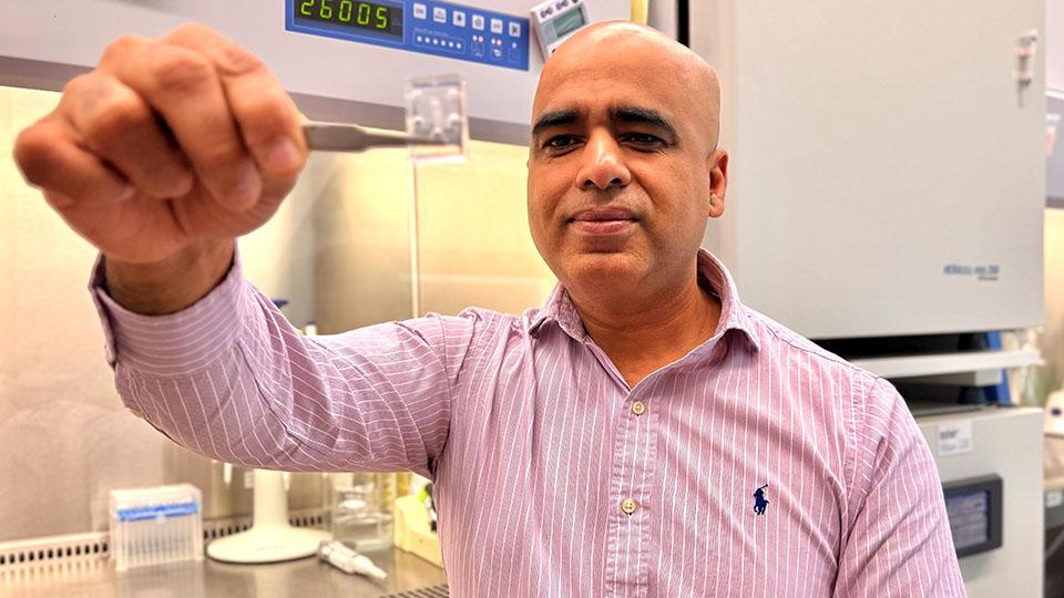 A man sits in a lab in front of a fume hood and uses tweezers to hold a plastic chip out toward the camera.