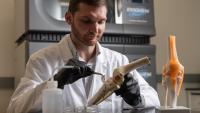 A man in a white lab coat and black gloves interacts with an implant and a knee model.
