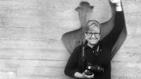 A woman holding a camera stands in front of a relief carving in a concrete wall mimicking its posture, with her arm raised over her head.