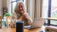 An elderly woman with short white hair smiles and looks at a smart speaker system.