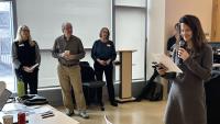 "A group of people stand near large windows in a bright indoor space, with one person holding a microphone and reading from a sheet of paper. Three individuals stand together near a lectern, some holding notes or drinks. In the foreground, a table holds laptops, papers, and a drink cup. The room has light-colored floors and tall window shades that diffuse the natural light."