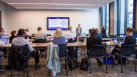 A photo shot from the back of a conference room with people sitting at conference tables while a person at the front of the room shows a presentation on a flat TV screen
