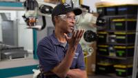 A man wearing a cap, safety goggles, and a navy shirt speaks in an industrial setting.